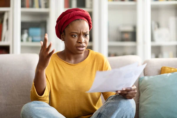 Furious angry young black woman in casual wearing red african turban sitting on couch, holding papers correspondence and gesturing, shocked with household bills, home interior, copy space