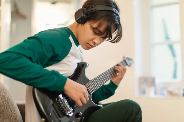 Asian Teen Guy Playing Electric Guitar Wearing Headphones Writing Song Sitting At Home. Boy Learning To Play Musical Instrument On Weekend. Teenagers Hobby And Music Talent. Selective Focus