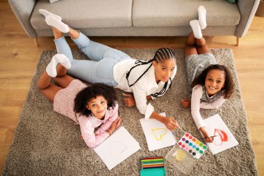 Mutlu siyah anne ve iki kızı, hafta sonları modern oturma odasında, kameraya gülümserken birlikte yerde yatıyorlar. Aile hobisi ve boş zaman. Görünümün Üzerinde Çekim