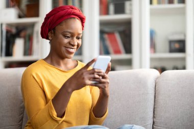 Olumlu, neşeli, Afrikalı türban takan genç bir kadın modern cep telefonu kullanıyor, flört uygulamasında erkeklerle sohbet ediyor, sosyal medyayı kontrol ediyor, blog okuyor, reklam için yer kopyalıyor.