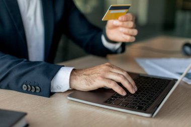 İş yerinde otururken ve online alışveriş yaparken kredi kartı ve laptop kullanan erkek girişimci, klavyeye odaklanıyor. İş adamı çevrimiçi bankacılık hizmeti üzerinden ödeme yapıyor