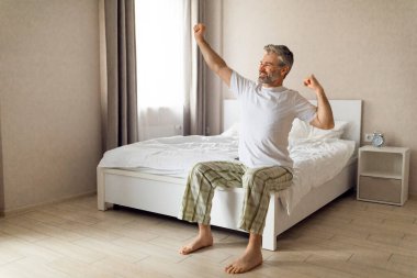 Neşeli, mutlu, yakışıklı, gri saçlı, orta yaşlı, yatakta oturan, kollarını uzatan, pencereye bakan, gülümseyen, güneşli günün tadını çıkaran, fotokopi çeken orta yaşlı bir adam. İyi uykular.