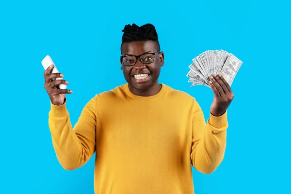 Excited black man holding dollar cash and smartphone in hands, happy young african american male celebrating online win and profit, standing isolated over blue studio background, copy space