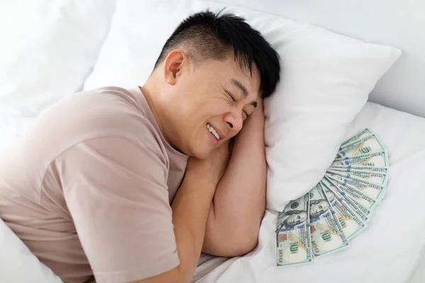 安详的亚洲人睡在床上 枕头底下藏着大量现金 微笑着 把他的钱藏在家里 特写照片 财务管理概念 — 图库照片
