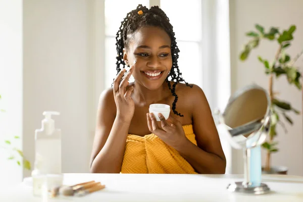 シャワーの後に顔のクリームを使用してタオルに包まれた陽気な黒の女性は メイクの準備ができて肌を作り 自宅でテーブルに座って 鏡を見て フリースペース — ストック写真