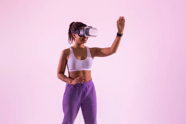 Excited black lady having fun with 3D technology, smiling cheerfully while exploring a virtual reality game, wearing a virtual reality headset over pink neon background