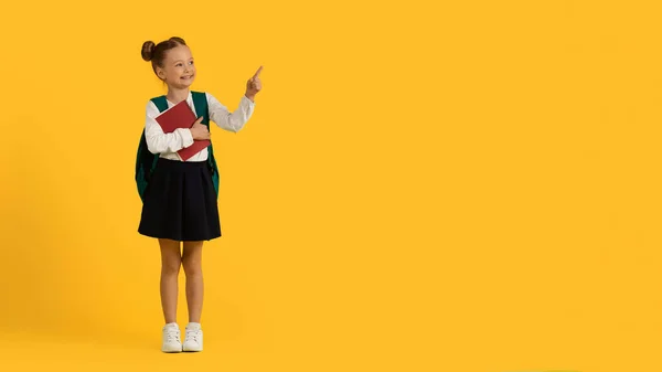 勉強広告 黄色の背景にコピースペースで脇を指してかわいい笑顔の女子高生 バックパックとワークブック付き幸せな小さな女性の子供広告のための無料の場所を示す パノラマ — ストック写真