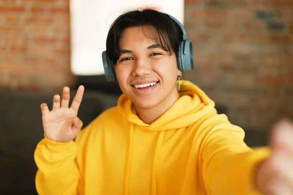 Hello. Happy Korean Teenager Guy Making Selfie Waving Hand Smiling To Camera Posing Wearing Headphones At Home. Young Blogger Making Video On Phone. Social Media And Gadgets. Selective Focus