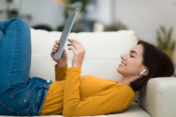 轻松地躺在沙发上 用现代数码垫和无线耳机 在互联网上观看电影或视频内容 复制空间的年轻女性 家庭娱乐 — 图库照片