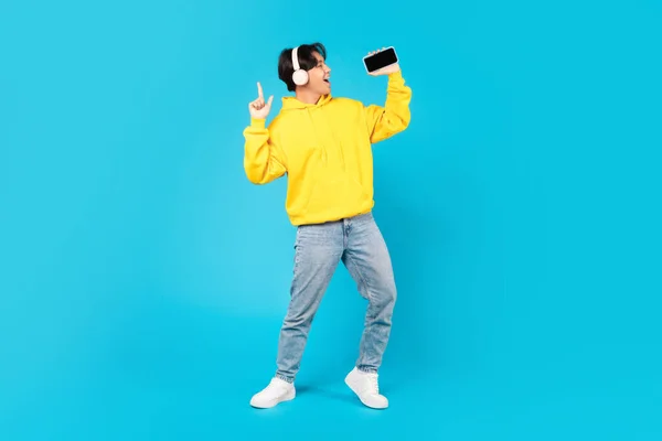 韓国の10代の男の子が青いスタジオの背景に立って楽しんでイヤホンを身に着けているマイクのようなスマートフォンを保持歌う 優れた音楽アプリケーション広告 フルレングスショット — ストック写真