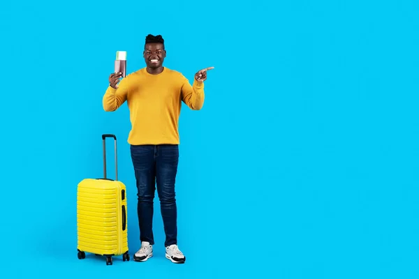 Happy african american man with suitcase and passport pointing aside at copy space, african american male traveller posing with luggage over blue studio background, full length, copy space