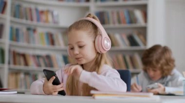 Okul çocukları ve modern teknolojiler. Kulaklık takmış şirin bir kız sınıfta akıllı telefondan sörf yapıyor, çocuk tatilde video oyunu oynuyor, boş yer var.