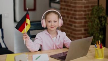 Yabancı erken eğitim. Kulaklık takan sevimli küçük bir kız dizüstü bilgisayarda çalışıyor, Almanya bayrağını sallıyor ve kameraya gülümsüyor, masa başında evde eğitimin keyfini çıkarıyor, ağır çekimde, boş alanda.