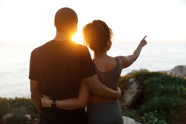 Young african american woman hugging man in sportswear, enjoy workout break, point finger at empty space in morning on rocks at ocean, outdoor. Relationship, health care, active lifestyle and sports