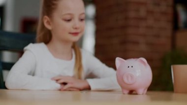 Çocuklar ve finansal okuryazarlık. Şirin küçük kız domuz kumbarasına para koyuyor ve el çırpıyor, gülüyor, evdeki ilk birikiminin tadını çıkarıyor, seçici odaklanma, boş alan.