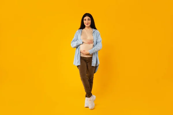 全长拍摄的快乐孕妇抱着肚子 对着镜头微笑 在黄色的工作室背景上摆姿势 温柔体贴地触摸着肚子 自由自在 — 图库照片