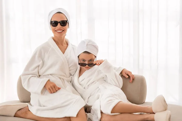 Positive Mom And Female Child In Bathrobes And Sunglasses Having Beauty Day At Home, Cheerful Mother And Daughter With Towels On Head Posing At Camera While Relaxing On Couch, Copy Space