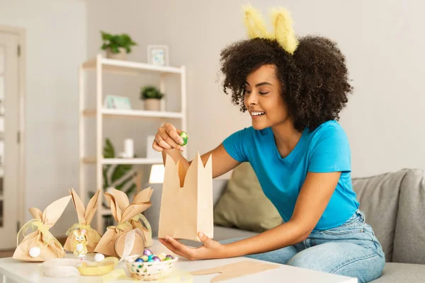 快乐的黑人妇女把复活节巧克力蛋包装在家里的纸袋里 快乐的年轻非洲裔美国女性穿着可爱的兔子耳朵做手工制作的礼品包装 享受节日庆祝 — 图库照片