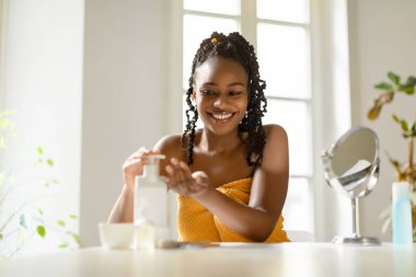 Mutlu Afrikalı Amerikalı kadın elinde şişeden nemlendirici vücut losyonu döküyor, havluya sarılmış, banyo üstüne yeni cilt ürünleri kullanıyor,