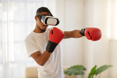 VR gözlüklü ve boks eldivenli genç bir sporcu. Görünmez birini yumrukluyor. Afro-Amerikan bir adam evde egzersiz yapıyor.