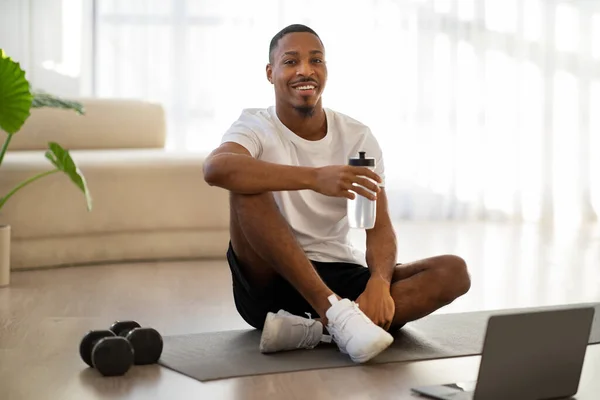 stock image Cheerful happy handsome sporty young black guy sitting on yoga mat in front of laptop, drinking water after workout online at home, copy space. Hydration during training concept