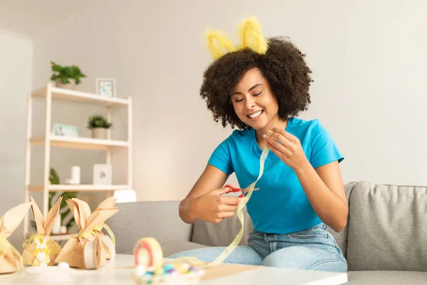 美丽的年轻黑人女性在家里做复活节装饰 快乐的非洲裔美国女人戴着兔子耳朵头带准备节日礼物包装 剪带和微笑 自由自在 — 图库照片