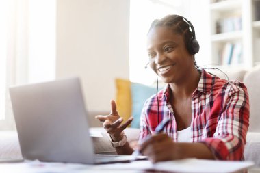 Neşeli, gülümseyen Afro-Amerikalı bir kadın bilgisayar başında oturmuş, kulaklık takıyor, notlar alıyor, online eğitime katılıyor ya da evdeki web sitelerini kopyalıyor.