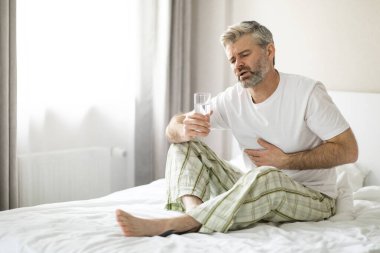 Mutsuz, hasta, orta yaşlı bir adam bir bardak suyla yatakta oturuyor ve karnına dokunuyor, sabahları mide ağrısı çekiyor, pijama giyiyor, iç mekan. Gastrobağırsak Hastalıkları, Sorunlar