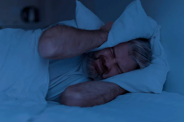 Closeup of angry mature man lying in bed and covering head ears with pillow at night, cannot sleep, suffering from noisy neighbours or snoring spouse partner