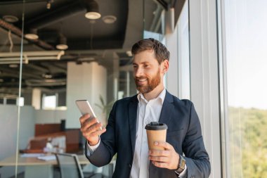 Takım elbiseli neşeli erkek yönetici cep telefonuyla bir bardak içkiyle sohbet ediyor, ofis içi pencerenin yanında duruyor, fotokopi odası. İş kumandası aygıtı