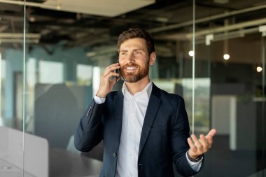 Kafkasyalı başarılı iş adamı akıllı telefonla konuşuyor, ofisin içinde duruyor, boş yer var. Pozitif erkek girişimci telefonda konuşuyor.