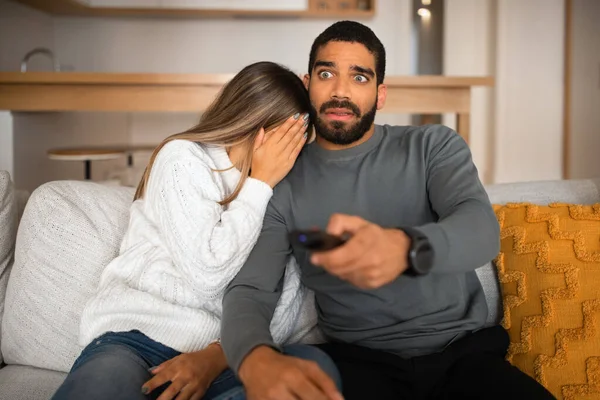 Shocked frightened millennial arab guy with beard, remote control and european woman close face, watch TV together on sofa in living room interior. Afraid, scared film at home and people emotions