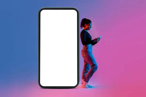 stock image Happy black lady in casual outfit leaning on huge cellphone with white blank screen, using smartphone, enjoying mobile app, mockup, colorful neon background, copy space