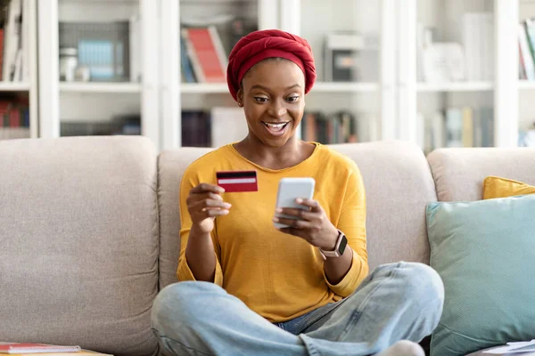 Online banking, Internet payment concept. Excited happy young african american woman in homewear customer sitting on couch at home, using red credit card and cell phone, copy space