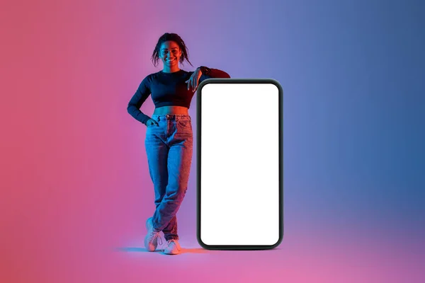 Happy black lady standing near giant cellphone with empty white screen in colorful neon light, mockup for mobile app, website, your ad design, template with copy space