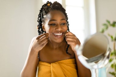Aynaya bakıp diş ipi kullanan, beyaz dişlerini temizleyen yatak odasının içindeki makyaj masasında oturan çekici siyahi kadın. Diş sağlığı konsepti