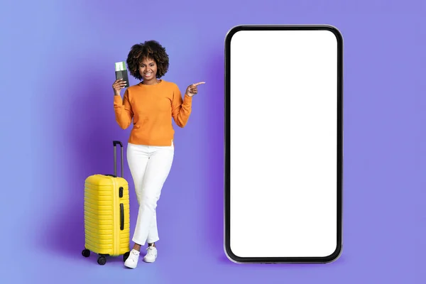 积极的黑人女性游客展示移动应用程序或在线提供游客 非洲女士与行李 门票指向大智能手机与白色屏幕 紫色背景 — 图库照片