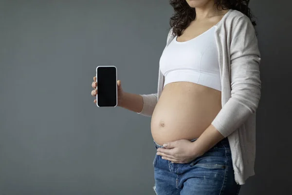 Unrecognizable Pregnant Woman Embracing Belly And Showing Blank Smartphone With Black Screen, Expectant Female Advertising Pregnancy App, Demonstrating Cellphone With Empty Screen, Mockup, Cropped