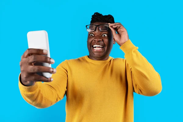 惊愕的黑人男性摘下眼镜 看着智能手机屏幕 惊讶的非洲裔美国年轻人站在蓝色背景下对网上拍卖的反应 — 图库照片