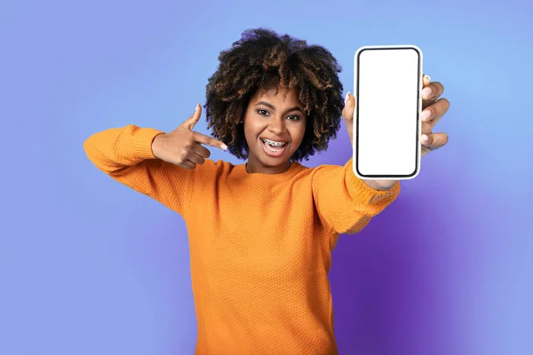一位年轻而快乐的非洲裔美国女士指着手中拿着白色空白屏幕的智能手机 面带微笑 在紫色的工作室背景下孤立无援 在网上给出了很好的报价 并进行了模仿 — 图库照片