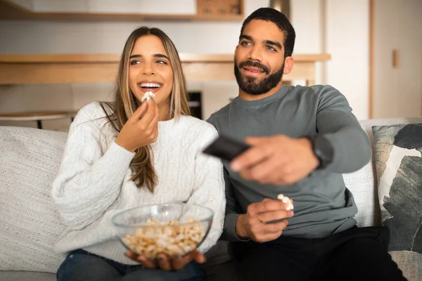 Glad millennial arab guy with beard and european female watch TV, eat popcorn, enjoy movie together on sofa in living room interior. Entertainment and fun at home snacks in free time, relationship