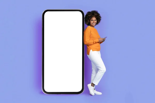 一个积极的 快乐的 漂亮的非洲年轻女士靠在巨大的手机上 屏幕为白色 使用智能手机 对着相机笑 背景为紫色 喜欢最新的应用程序 — 图库照片