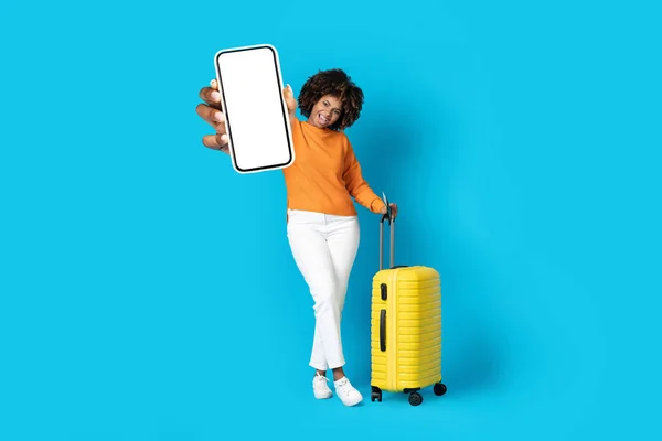 幸せな正の若いアフリカ系アメリカ人女性でカジュアルな観光客で黄色の荷物をスマートフォンに白い空白の画面で表示し 旅行アプリ 青の背景 モックアップ コピースペースをお勧めします — ストック写真