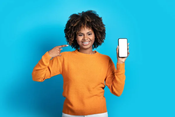 快乐而可爱的漂亮的非洲裔美国女士指着手里拿着白色空白屏幕的智能手机微笑着 在蓝色的工作室背景下孤立无援 在网上给出了很好的报价 模仿着 — 图库照片