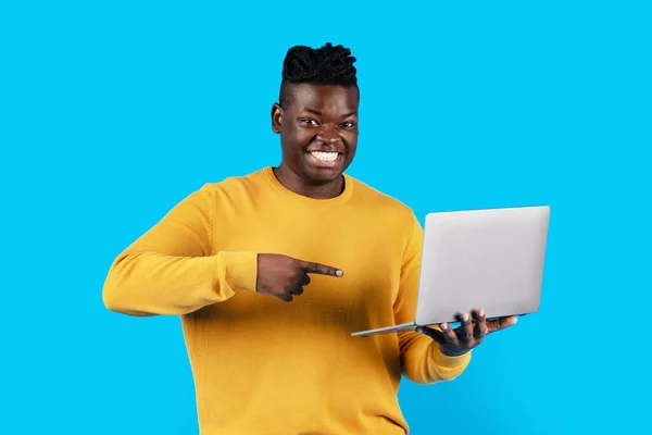 Portrait Of Cheerful Young Black Man Pointing At Laptop In His Hands, Happy Millennial African American Male Demonstrating Computer, Recommending New Website While Standing Over Blue Background