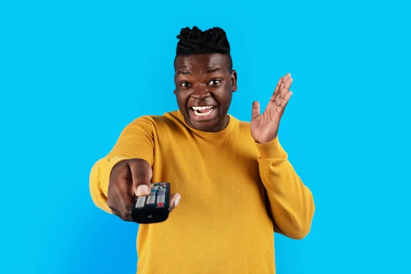 Excited black man holding remote control in hand and pointing it at camera, joyful african american guy watching tv, switching channels while standing over blue studio background, copy space