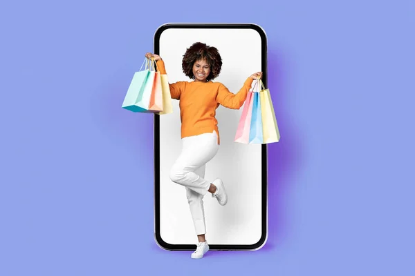 漂亮而快乐的年轻黑人女人 穿着休闲装 浓密的头发和购物狂一起跳到大手机旁边 白色空白屏幕 微笑着 提着购物袋 紫色背景 复制空间 — 图库照片