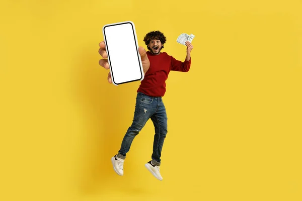 Online bet, gambling, trading on Internet, giveaway. Happy young indian guy jumping in the air with phone and cash in his hands, showing smartphone with white blank screen, mockup, yellow background