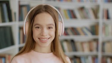 Mutlu öğrenci hayatı. Oldukça güzel bir genç bayan öğrenci kablosuz kulaklık takıyor kameraya gülümsüyor, kütüphanenin içinde boş bir alanda poz veriyor.