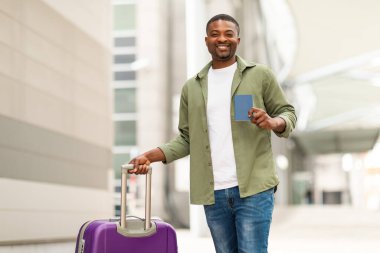 Ucuz biletler. Pasaportunu gösteren mutlu Afrikalı Amerikalı adam seyahat çantasıyla havaalanı terminalindeki kameraya gülümsüyor. Tatil Turu Teklif Konsepti